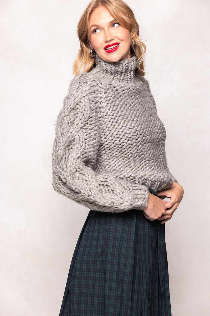 A woman stands sideways and smiles, one hand on her waist, wearing the Allegra Sweater in Gray by Ivy City Co—a chunky wool cable-knit turtleneck paired with a dark green plaid skirt for a cozy, stylish look that’s also maternity friendly.