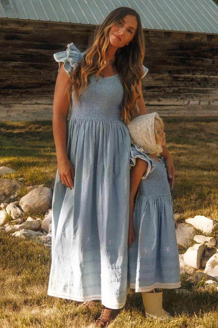 A woman and a young girl, both wearing matching Ivy City Co Hattie Ankle Length Dresses in Embroidered Chambray with smocked bodices, stand together outside on grass with rocks nearby and a rustic wooden building in the background. These dresses are maternity friendly, making them perfect for every stage of life.