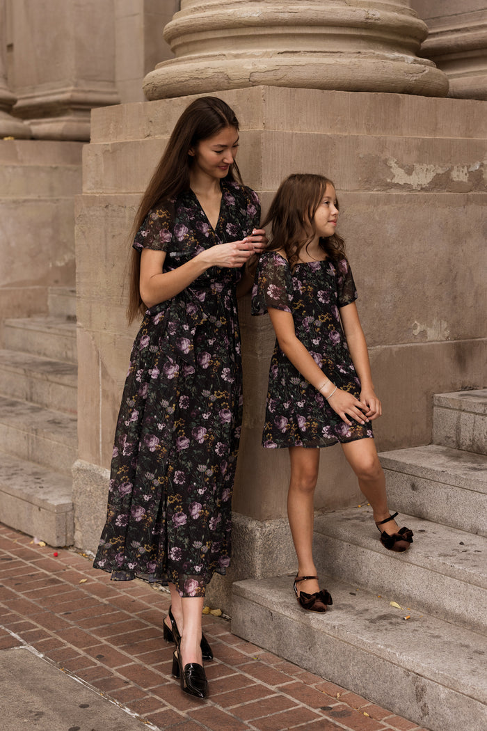 A woman and a girl, both dressed in matching Lyla Dresses in Belladonna Bloom by Ivy City Co, stand on stone steps beside a large building. The nursing-friendly design of the dress offers both comfort and style as the woman checks her phone while the girl stands next to her, gazing off into the distance.