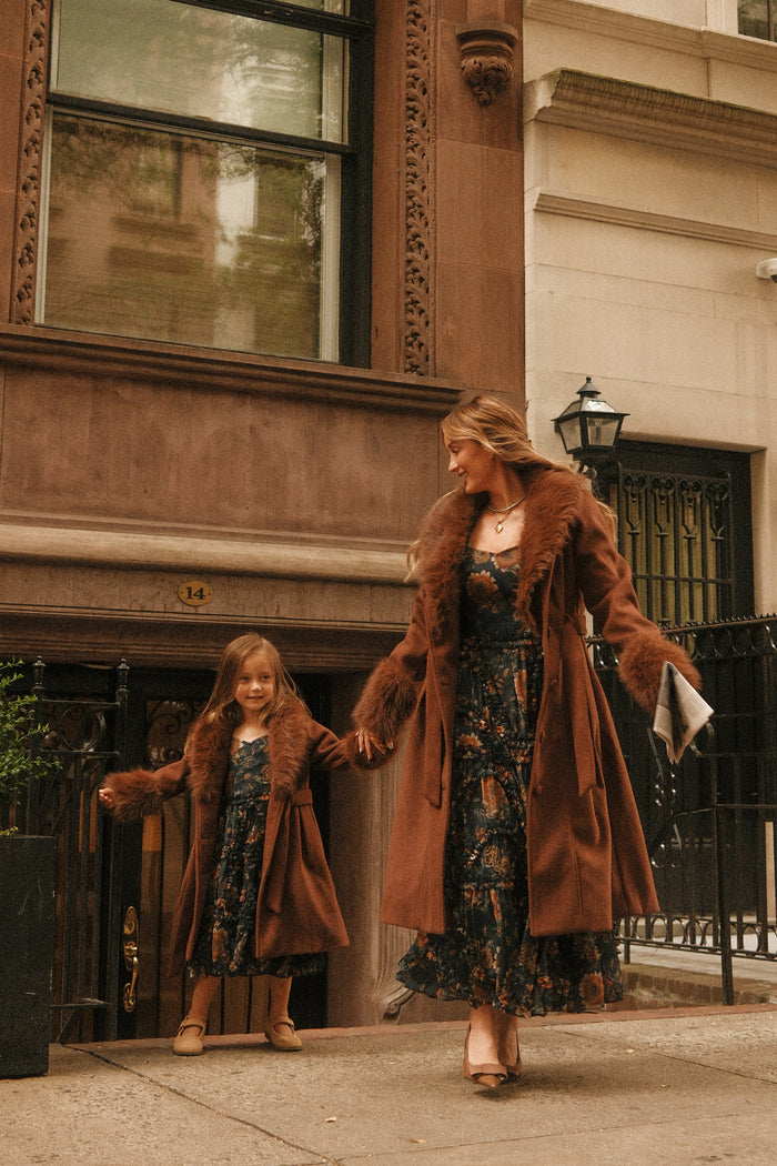A woman and a young girl, both dressed in matching brown coats with fur trim and floral dresses, hold hands and smile as they stand on a city sidewalk in front of a brownstone building. The girl’s cozy outfit highlights the charming Mini Pookie Coat in Warm Cocoa by Ivy City Co—an ideal girls’ fall coat for crisp days around the city.