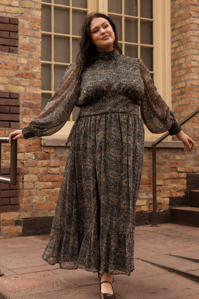 A woman wearing the Ivy City Co Payton Dress in Evergreen Paisley, featuring sheer sleeves and a flowing maxi silhouette, stands outdoors smiling in front of a brick wall and large window.