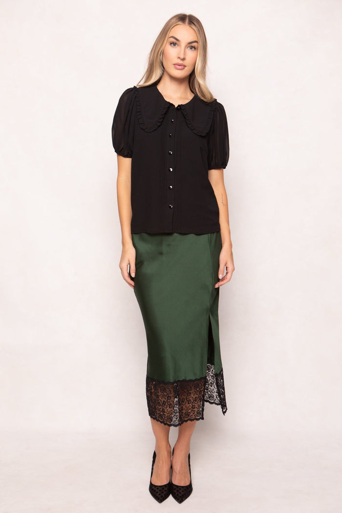 A woman with long blonde hair and a neutral expression stands against a plain background, wearing the Ivy City Co Andrea Blouse in black chiffon, styled with a green satin skirt featuring black lace trim and paired with black high heels.