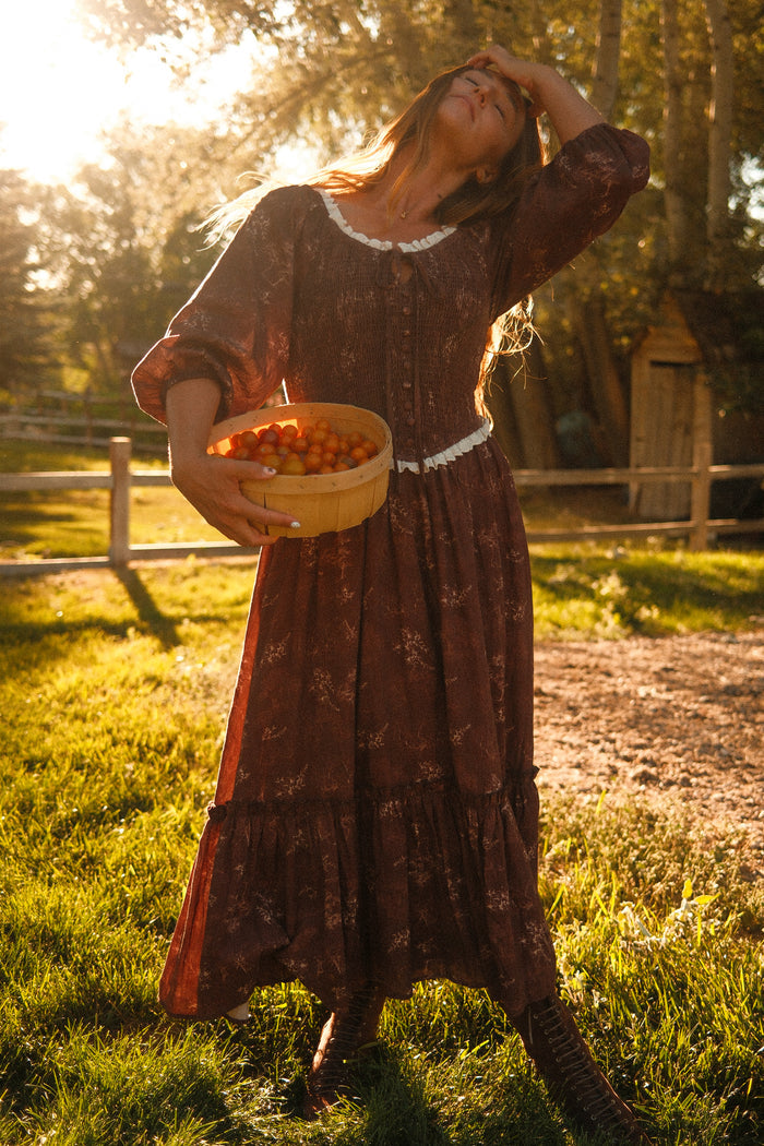 Basked in sunlight in a grassy yard with trees and a wooden fence, a woman stands wearing the Ivy City Co Asha Dress in Warm Cocoa Valencia Toile—a long, flowy cotton dress offering both style and comfort that’s even maternity friendly. She tilts her head back, touching her forehead while holding a basket of yellow-orange tomatoes.