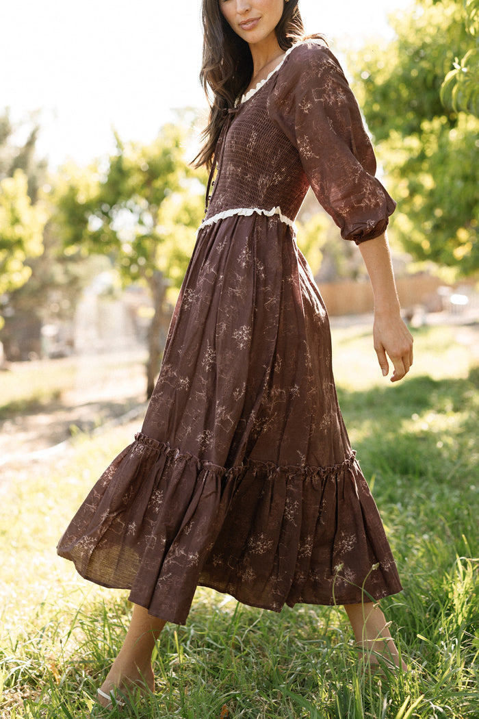 A woman walks barefoot on grass in a sunlit outdoor setting with trees in the background, wearing Ivy City Co’s Asha Dress in Warm Cocoa Valencia Toile—a long, brown floral cotton dress featuring puffed sleeves. This maternity-friendly style provides comfort and effortless elegance for warm, leisurely days.