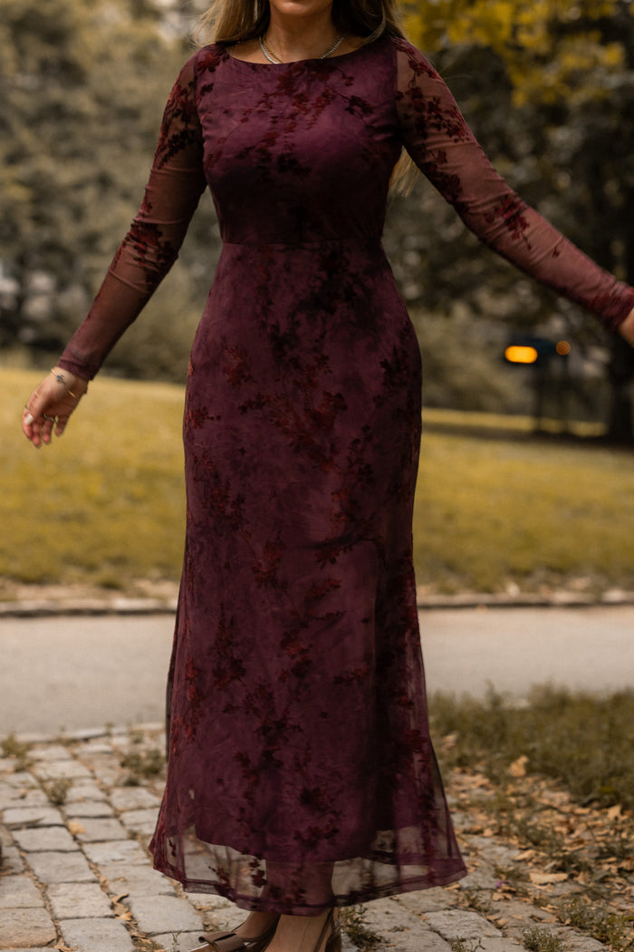A woman stands outdoors wearing the Ivy City Co Blair Dress in Maroon Burnout Floral. The dress is long-sleeved, floor-length, and features a sheer overlay. Grass, trees, and a paved walkway appear in the background. Her face is not visible.