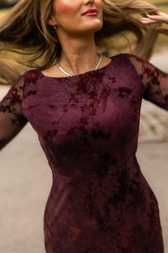 Outdoors, a woman with long hair in motion twirls while wearing the Ivy City Co Blair Dress in Maroon Burnout Floral, which features sheer sleeves and is paired with a necklace. Her face is partially visible.