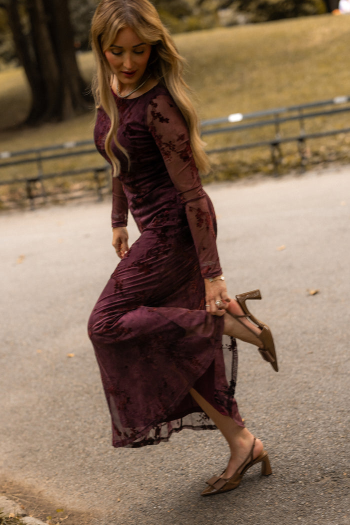 An Ivy City Co Blair Dress in Maroon Burnout Floral is worn by a woman standing outdoors on a path, lifting one foot to adjust her brown heels, with grass and trees in the background.