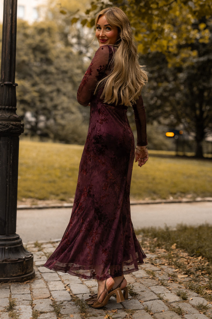 A woman wearing the Ivy City Co Blair Dress in Maroon Burnout Floral, featuring sheer sleeves and a long silhouette, stands on a cobblestone path outdoors. She looks back over her shoulder with a smile, surrounded by trees and grass in the background.