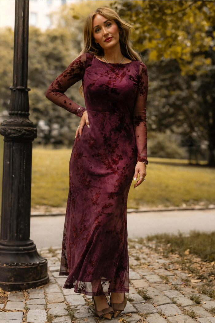 A woman wearing the long, fitted Blair Dress in Maroon Burnout Floral by Ivy City Co stands on a cobblestone path near a black lamppost in a park, the deep maroon hue and sheer sleeves of her maxi evening dress mirroring the elegance of the surrounding trees and grass.