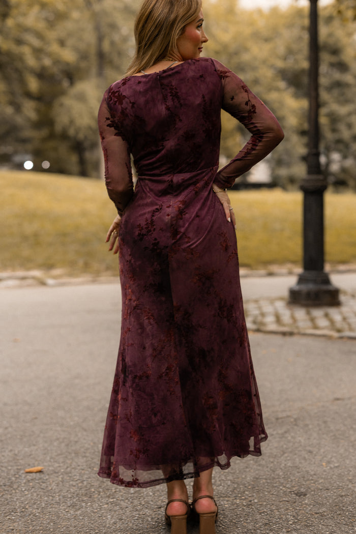 A woman stands outdoors on a path, facing away from the camera, wearing the Ivy City Co Blair Dress in Maroon Burnout Floral—an elegant maxi evening dress featuring sheer long sleeves—paired with pink heels. Trees and grass are visible in the background.