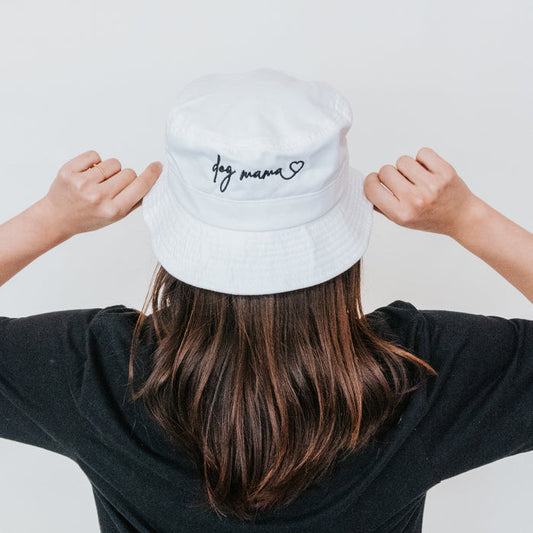 A person with long brown hair in a black shirt faces away, pointing at the Sassy Woof Bucket Hat - Dog Mama, a white bucket hat embroidered with “dog mama” and a heart.