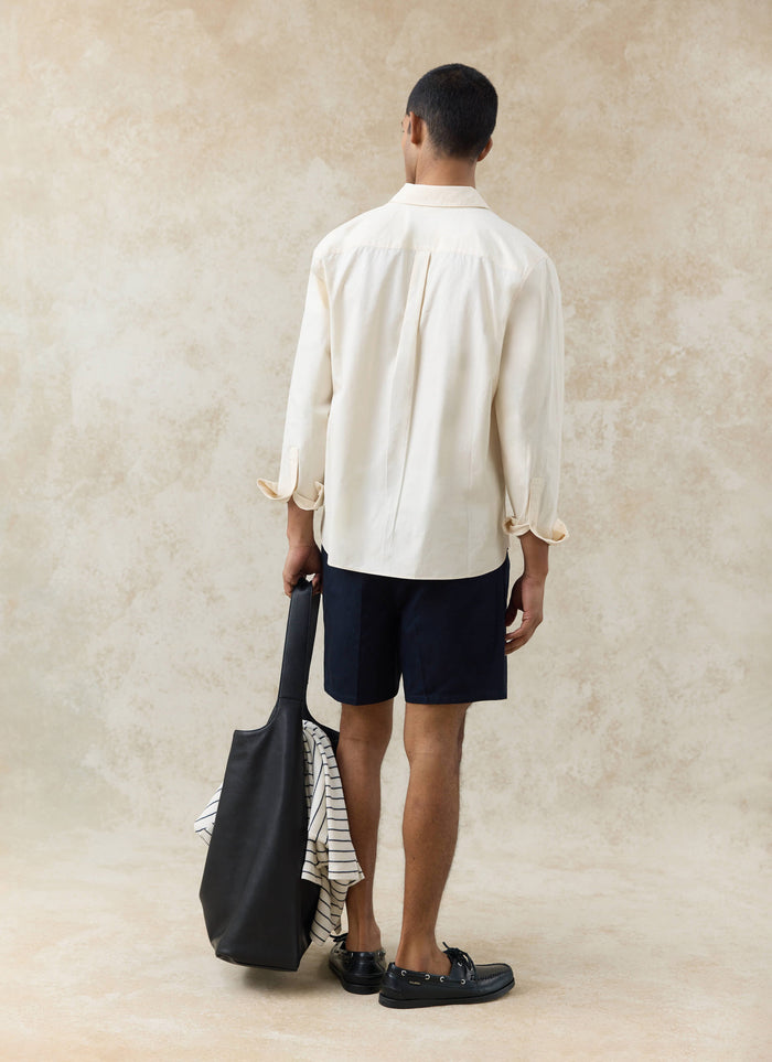 Rear view of the Percival Dali Relaxed Poplin Shirt in cream cotton, styled with navy shorts and a large black tote bag.