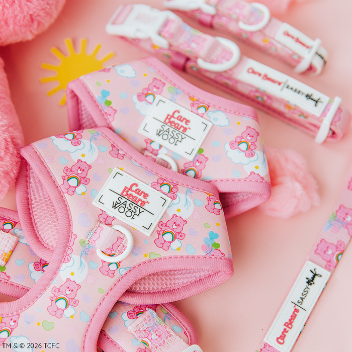 The Sassy Woof Dog Adjustable Harness - Care Bears™ Cheer Bear features cheerful bear faces, rainbows, and clouds on a soft pink background, with plush toys and a yellow sun completing this vibrant pet accessory.