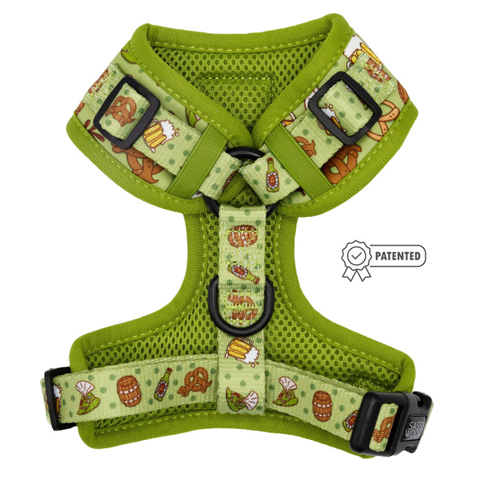 The Sassy Woof Dog Adjustable Harness - Wurst Behavior is a green mesh no-pull harness with cartoon pretzels, beer mugs, and sausages. It features black buckles, metal rings, and a PATENTED badge on the right side.