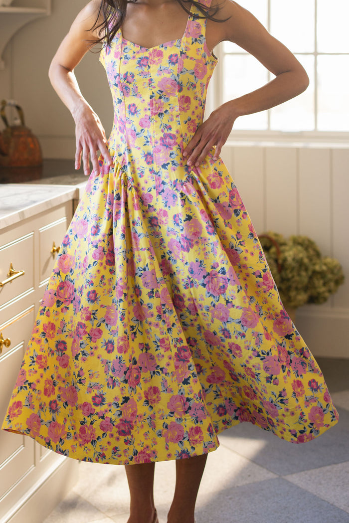 A woman stands in a sunlit kitchen wearing the Ivy City Co Edith Dress in As You Dream Floral, a sleeveless yellow dress with pink and purple blooms, featuring a fitted bodice and flared midi skirt as she rests her hands on her waist.