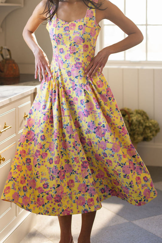 A woman stands in a sunlit kitchen wearing the Ivy City Co Edith Dress in As You Dream Floral, a sleeveless yellow dress with pink and purple blooms, featuring a fitted bodice and flared midi skirt as she rests her hands on her waist.