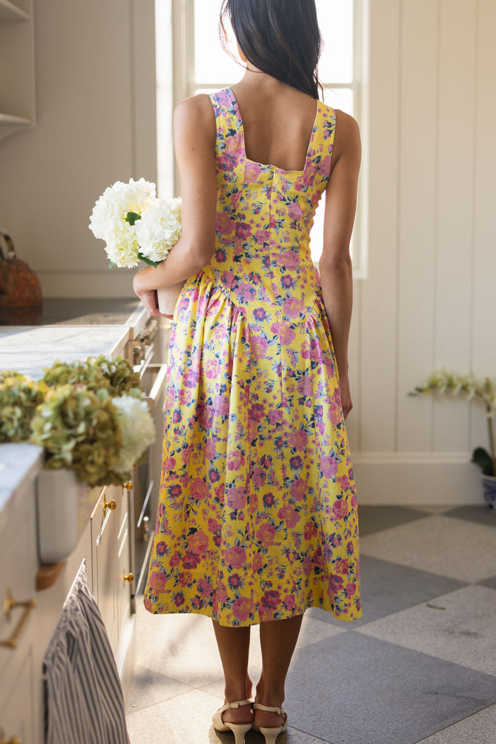A woman, back turned to the camera in a sunlit kitchen, holds white flowers while wearing Ivy City Co’s Edith Dress in As You Dream Floral—a sleeveless yellow and pink floral spring dress—with sandals as light streams onto blooms on the counter.