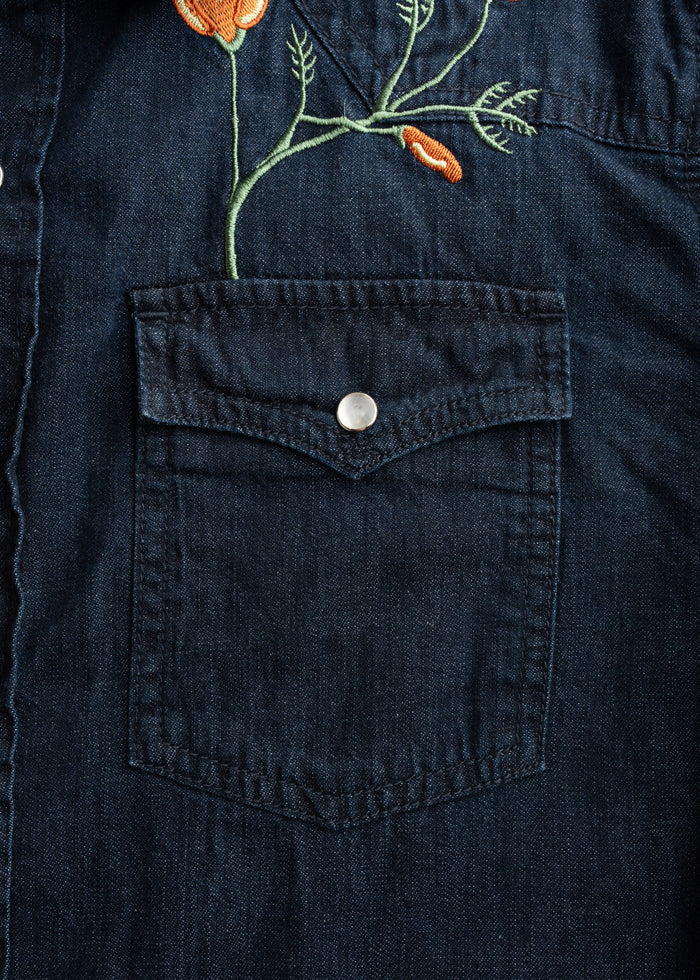 Denim shirt pocket with orange floral embroidery, featuring a button closure. The design adds a stylish touch to the fabric.