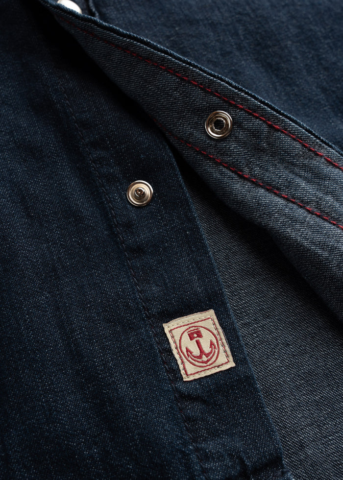 Close-up of a denim jacket with red stitching, metal snaps, and a small anchor patch, showcasing detailed craftsmanship.