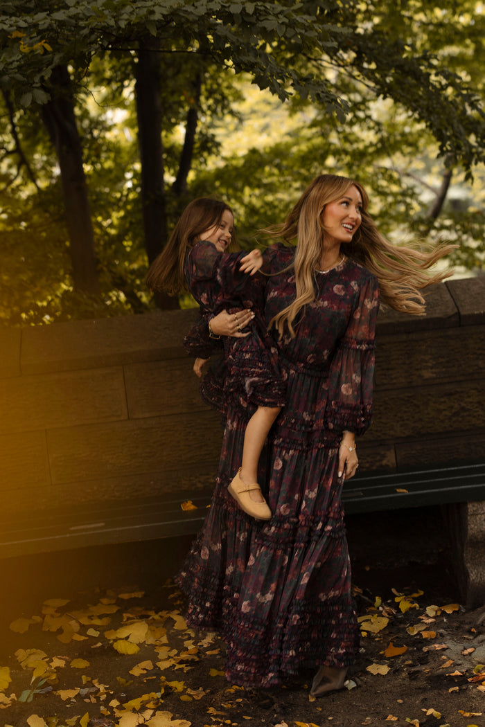 A woman wearing the Ivy City Co Geneva Dress in Aubergine Resplendent Bloom holds a smiling young girl, also dressed in matching Ivy City Co attire, outdoors beside a bench and surrounded by trees with fallen yellow leaves.