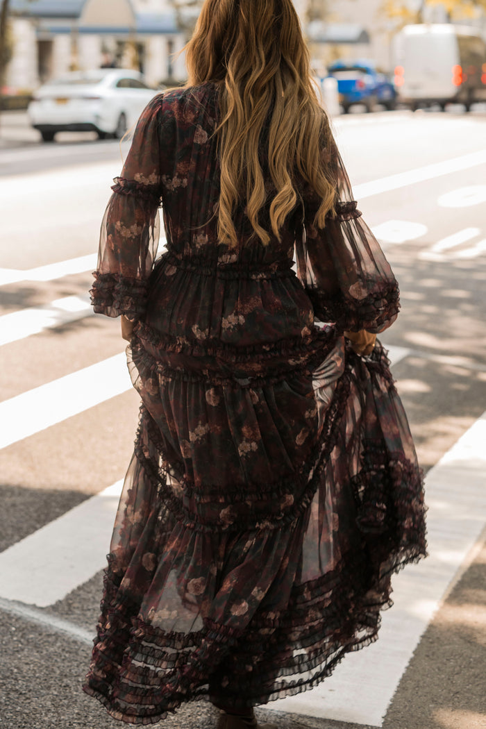 A person with long wavy hair wearing the Geneva Dress in Aubergine Resplendent Bloom by Ivy City Co crosses a street at a crosswalk on a sunny day.