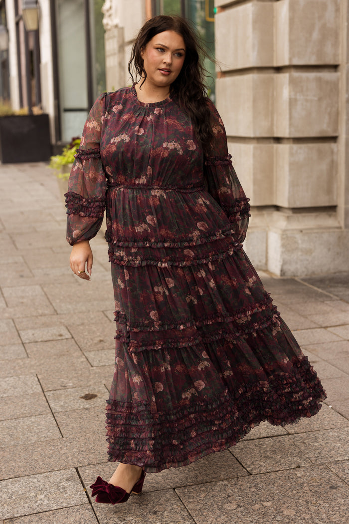 A woman wearing the Ivy City Co Geneva Dress in Aubergine Resplendent Bloom, featuring elegant balloon sleeves, pairs it with red shoes as she walks along a city sidewalk near a stone building, glancing slightly to her left.