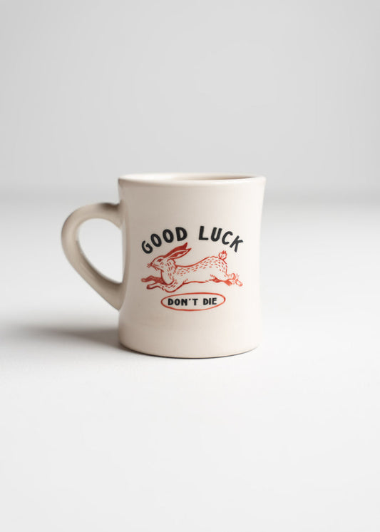 A white mug with a red rabbit design and the words "GOOD LUCK DON'T DIE" in bold letters on a plain background.