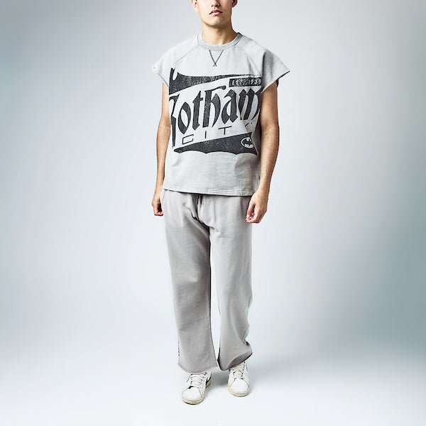 Man in a Sleeveless light gray sweatshirt featuring a large vintage-style graphic of "Gotham City" with a bat silhouette and the text "Est. 1939" in distressed black print. A small Batman logo is also present at the bottom right of the design Light gray sweatpants with an elastic waistband and black drawstring, featuring a small black Batman logo near the left pocket and a vertical "Gotham City" graphic printed in distressed black text along the lower left leg.