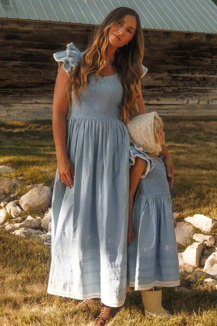 A woman and young child stand near a rustic barn, both wearing the Hattie Ankle Length Dress in Embroidered Chambray by Ivy City Co. These coordinated blue dresses feature ruffle sleeves, a smocked bodice, and white embroidered hems. Elegantly maternity friendly, this style is perfect for mothers and their little ones to match outdoors.