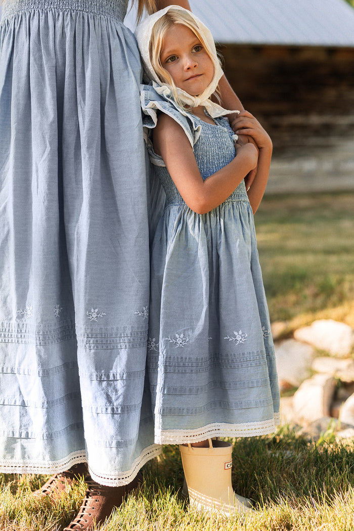 A young blonde girl in the Ivy City Co Hattie Ankle Length Dress in Embroidered Chambray, featuring a smocked bodice, hugs an adult wearing a matching dress. They stand on grass near a rustic building. The girl, looking thoughtful and wearing beige rain boots, embodies the charm of this maternity-friendly style—perfect for creating memories together.