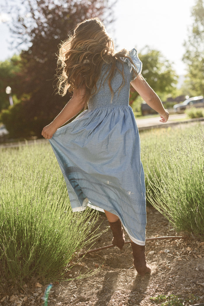 A girl with long wavy hair wearing the Ivy City Co Hattie Ankle Length Dress in Embroidered Chambray, which is maternity friendly, runs down a path through tall grass on a sunny day. She holds up the skirt with its smocked bodice and wears brown boots.