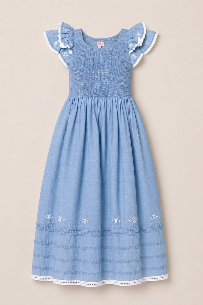 The Ivy City Co Hattie Ankle Length Dress in Embroidered Chambray is a light blue, sleeveless dress for girls, featuring ruffled lace-trim sleeves, a smocked bodice for comfort and style, and delicate embroidered flowers along the hem. Displayed on a neutral background, this charming dress is maternity friendly, making it a versatile addition to any wardrobe.
