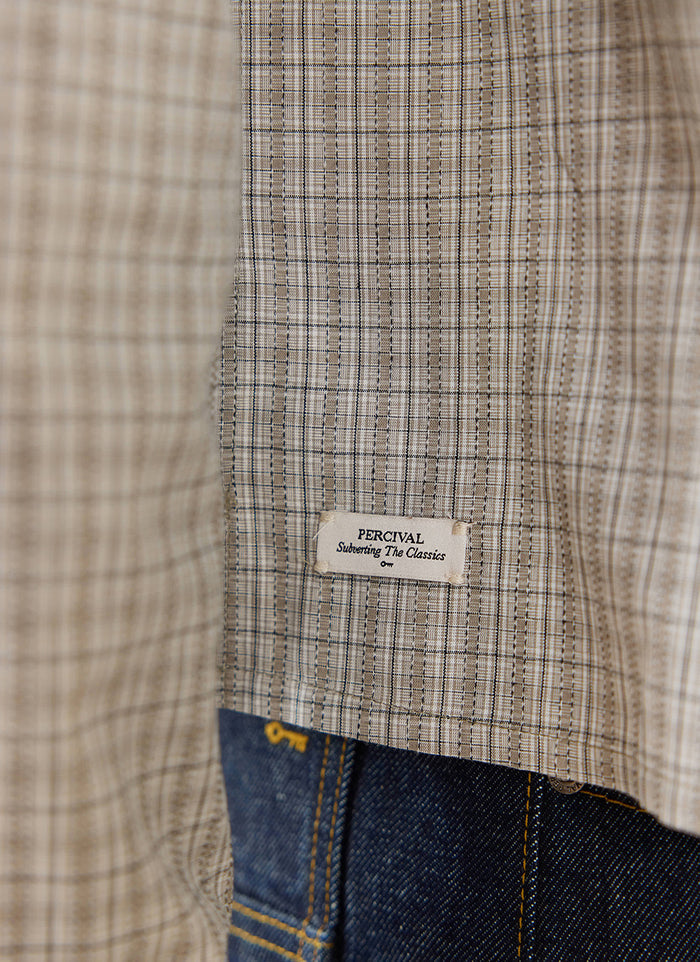 A close-up of the cream check Hinsley shirt by Percival, highlighting the branded hem tag and dark denim jeans.