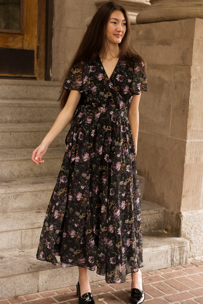 A woman in the Ivy City Co Lyla Dress in Belladonna Bloom stands on stone steps, smiling slightly. She has long brown hair and wears black shoes. Set against a wooden door and tan stone wall, this nursing-friendly maxi dress combines elegance with practicality.