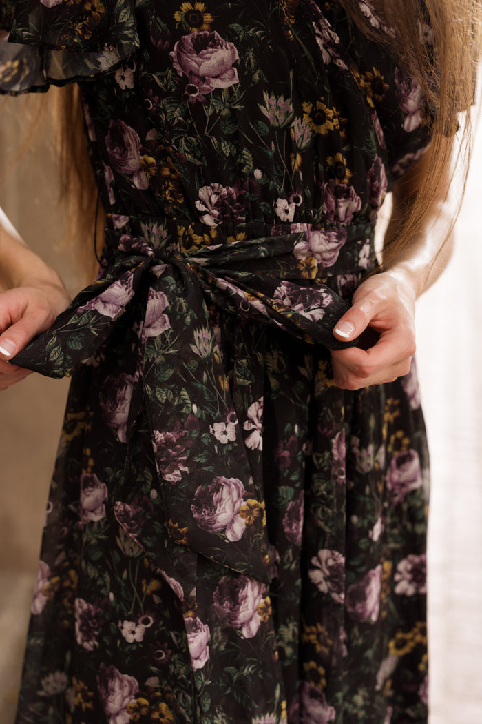 A person wearing the Lyla Dress in Belladonna Bloom by Ivy City Co ties a fabric belt at their waist. Only their torso and hands are visible, with long brown hair draped over one shoulder, highlighting this elegant maxi dress that is both stylish and nursing friendly.
