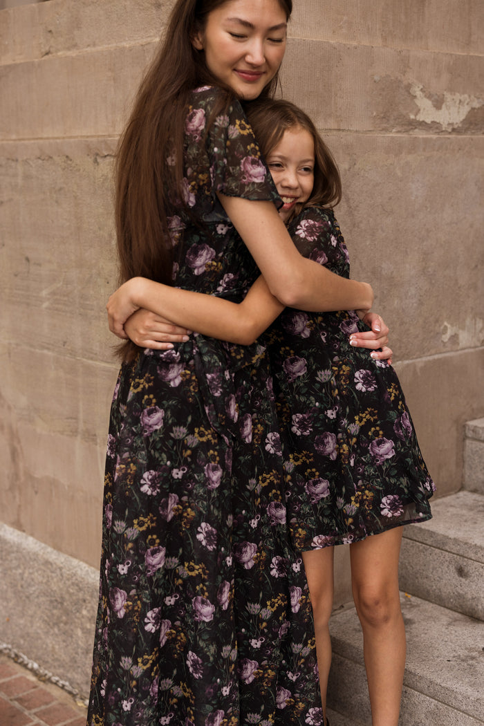 Two people, one adult and one child, wearing matching Lyla Dresses in Belladonna Bloom by Ivy City Co, hug each other and smile while standing outside near stone steps and a brick wall.