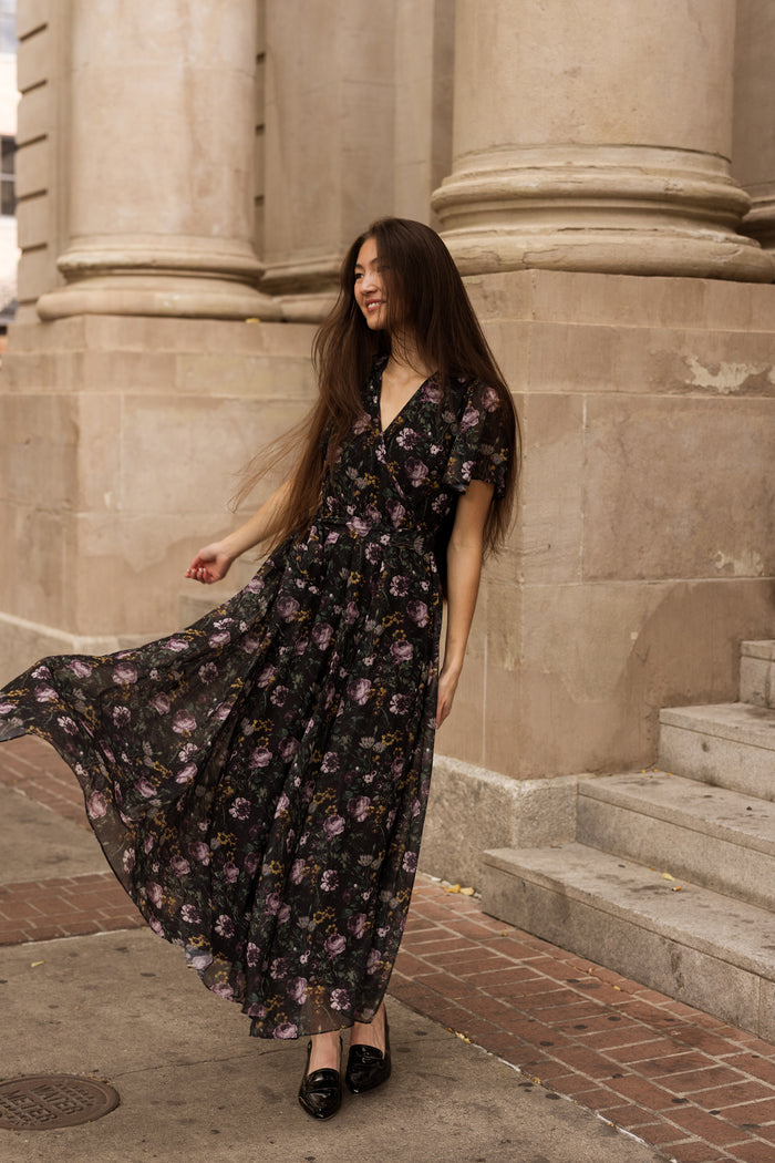 Wearing the Ivy City Co Lyla Dress in Belladonna Bloom, a woman stands on a sidewalk near stone columns and steps, smiling as her nursing-friendly maxi dress catches the breeze.