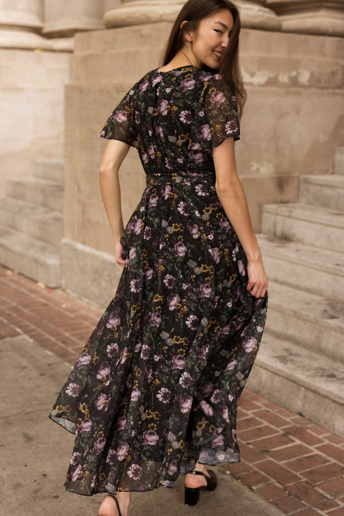 A woman walks outdoors near stone steps, looking back with a smile in her long, flowy Lyla Dress in Belladonna Bloom by Ivy City Co. The black maxi dress features elegant purple floral patterns and offers a nursing-friendly design for added comfort as it moves gracefully with each step.