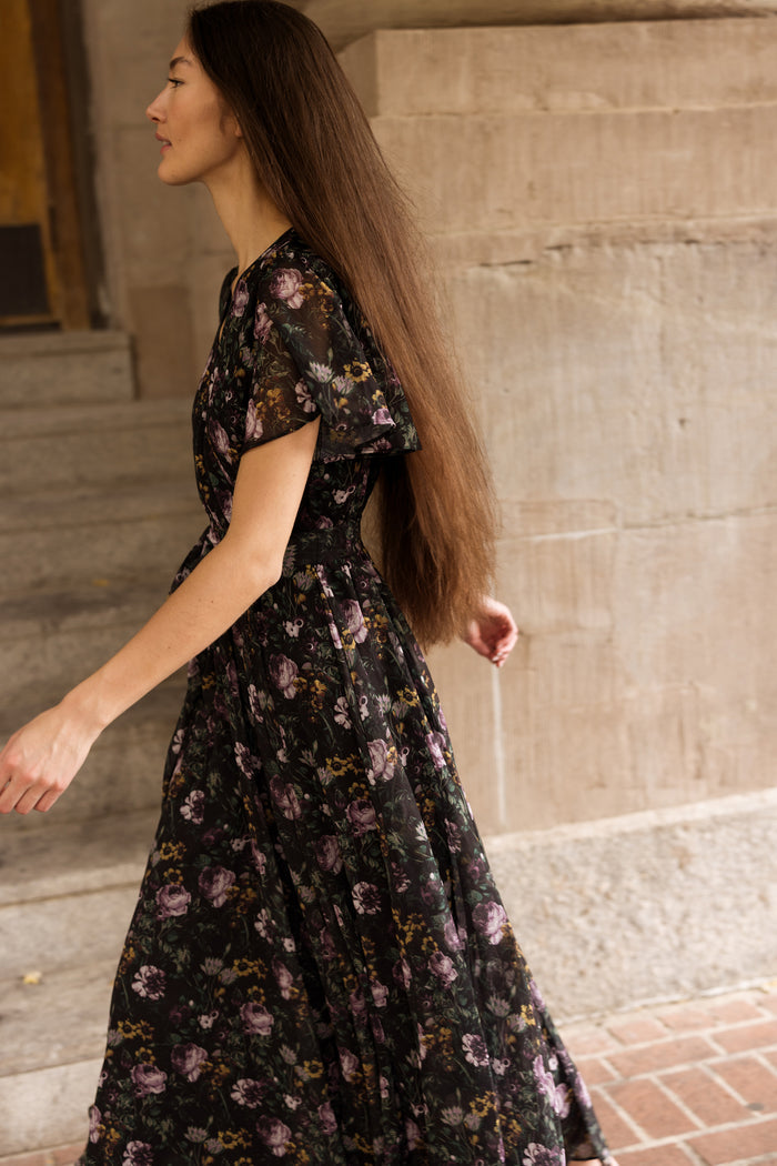 A woman with long brown hair, wearing the Ivy City Co Lyla Dress in Belladonna Bloom, walks past a stone wall on a brick sidewalk, photographed from the side.