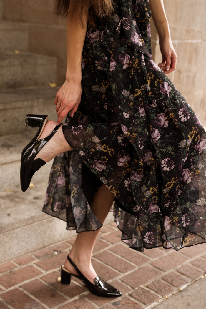 Wearing the Ivy City Co Lyla Dress in Belladonna Bloom, a person adjusts their black heeled shoe while standing on a brick and stone staircase.