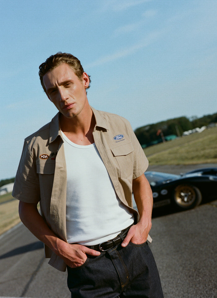 A model wears the beige Ford x Percival embroidered garage shirt with dark denim jeans in a vintage racing setting.