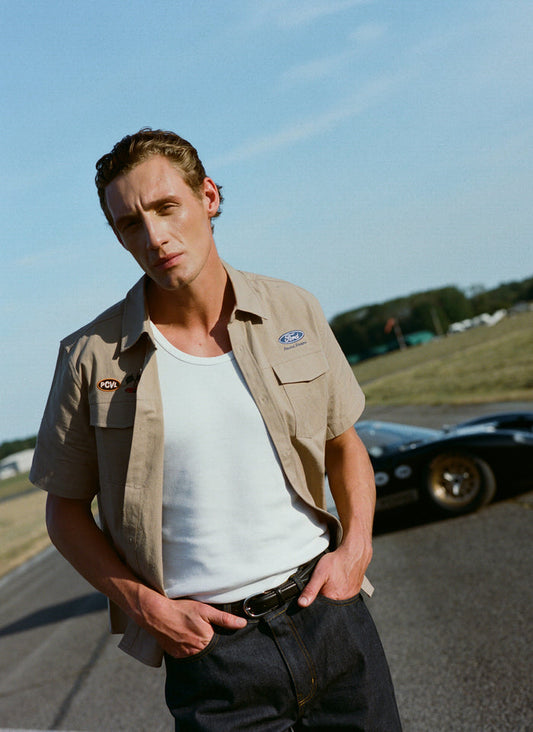 A model wears the beige Ford x Percival embroidered garage shirt with dark denim jeans in a vintage racing setting.