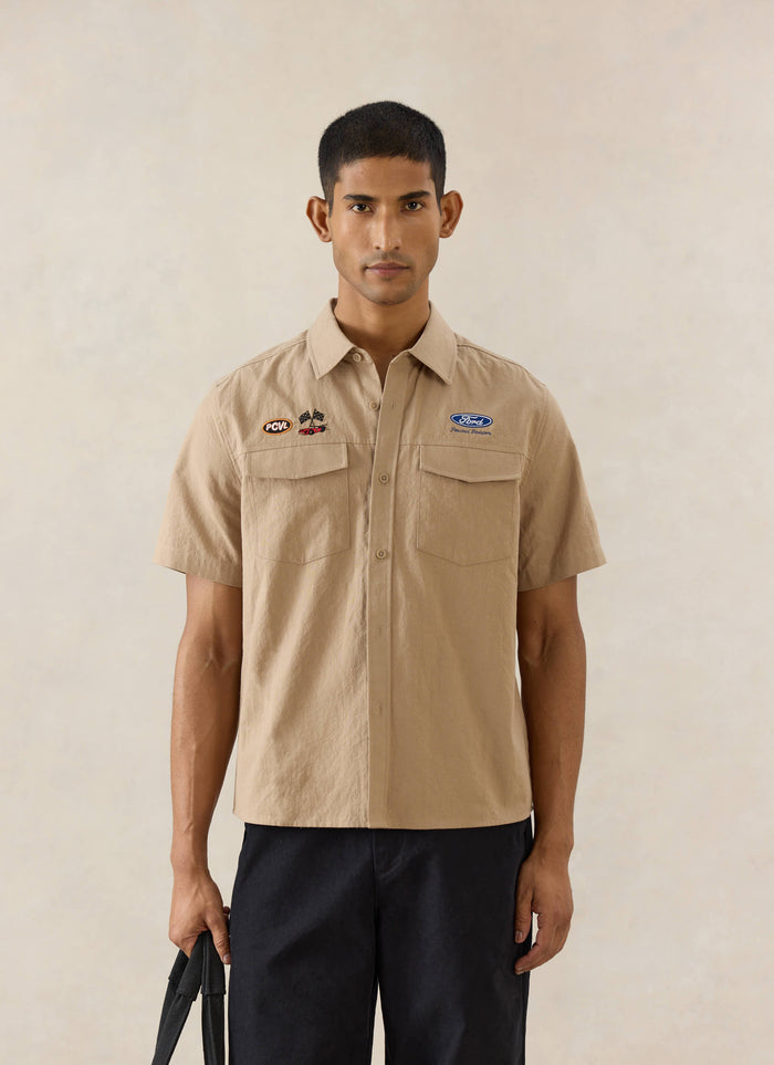 A model wears the beige Percival x Ford Miles Garage shirt with embroidered badges, styled with raw heritage denim jeans.