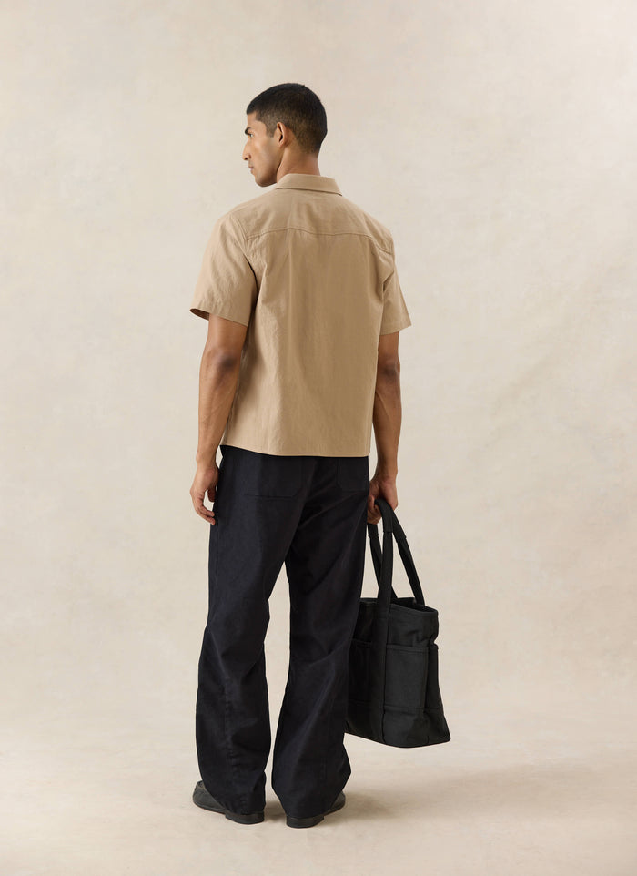 A model wears the beige Ford x Percival garage shirt from behind, styled with dark straight leg heritage denim jeans.