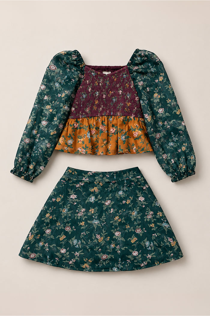 The Mini August Set in Elysian Vine by Ivy City Co features a two-piece design with a maroon long-sleeve floral top showcasing stylish balloon sleeves, a smocked bodice, and an orange floral panel, paired with a coordinating green floral skirt.