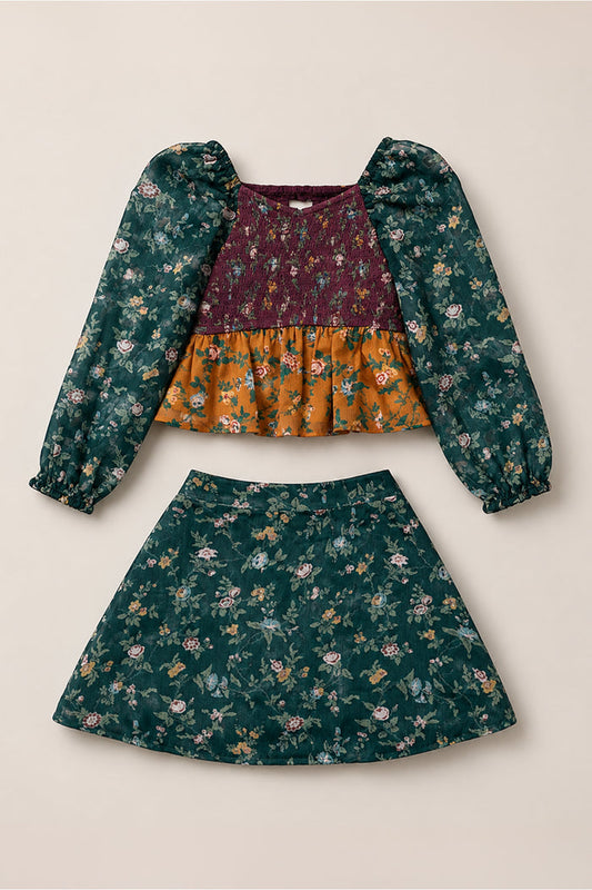 The Mini August Set in Elysian Vine by Ivy City Co features a two-piece design with a maroon long-sleeve floral top showcasing stylish balloon sleeves, a smocked bodice, and an orange floral panel, paired with a coordinating green floral skirt.