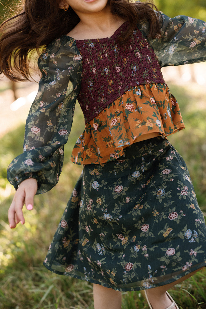 A young girl wearing the Ivy City Co Mini August Set in Elysian Vine, featuring balloon sleeves and a smocked bodice, spins outdoors on the grass with her arms outstretched and hair flowing, her face partially out of frame.