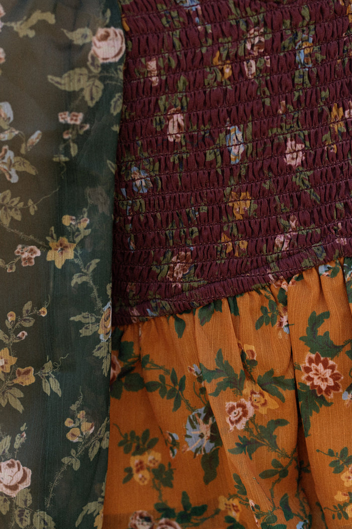Close-up of the Mini August Set in Elysian Vine by Ivy City Co—this floral dress showcases a smocked burgundy bodice, golden-orange skirt, and delicate balloon sleeves, displayed beside a dark green floral fabric.
