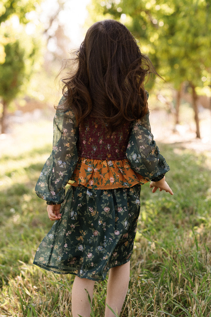 A young child with long brown hair, wearing the Ivy City Co Mini August Set in Elysian Vine—a green floral outfit with a smocked bodice—stands on grass, pointing to the side and facing away in a sunlit outdoor setting with trees in the background.