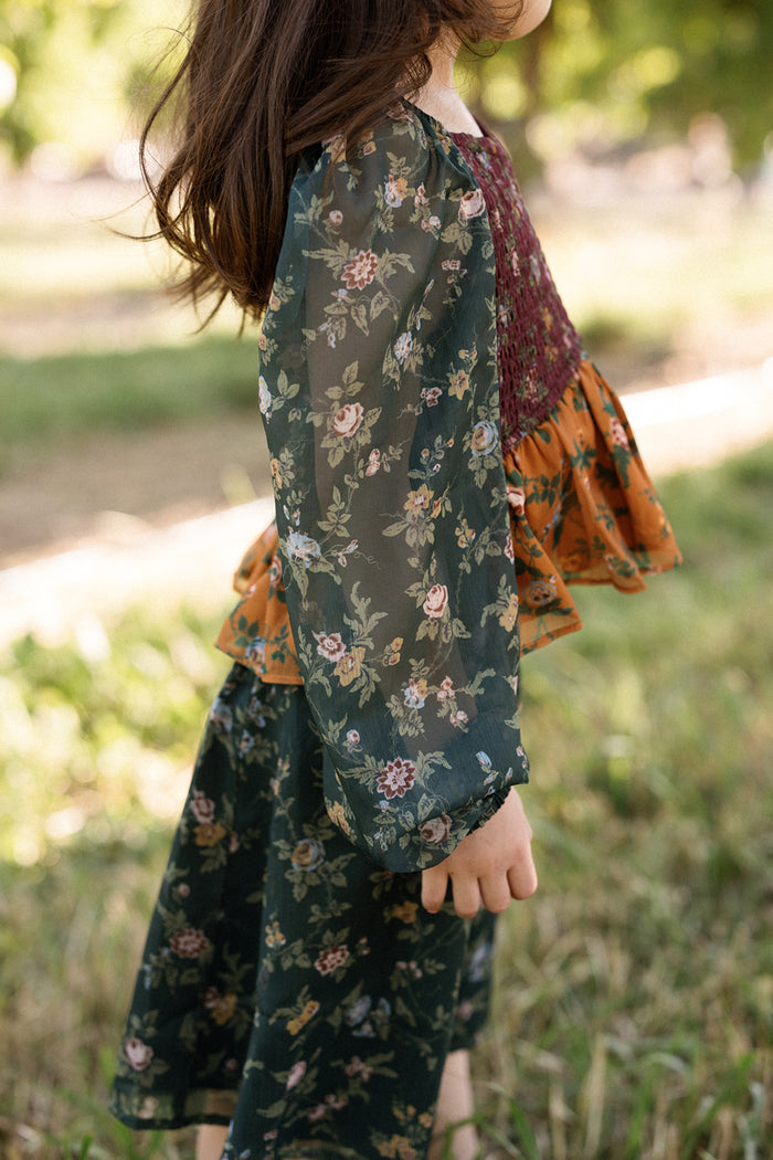 A child stands outside on grass, facing sideways, wearing the Ivy City Co Mini August Set in Elysian Vine—a long-sleeved, dark green floral dress with sheer balloon sleeves and an orange smocked bodice.